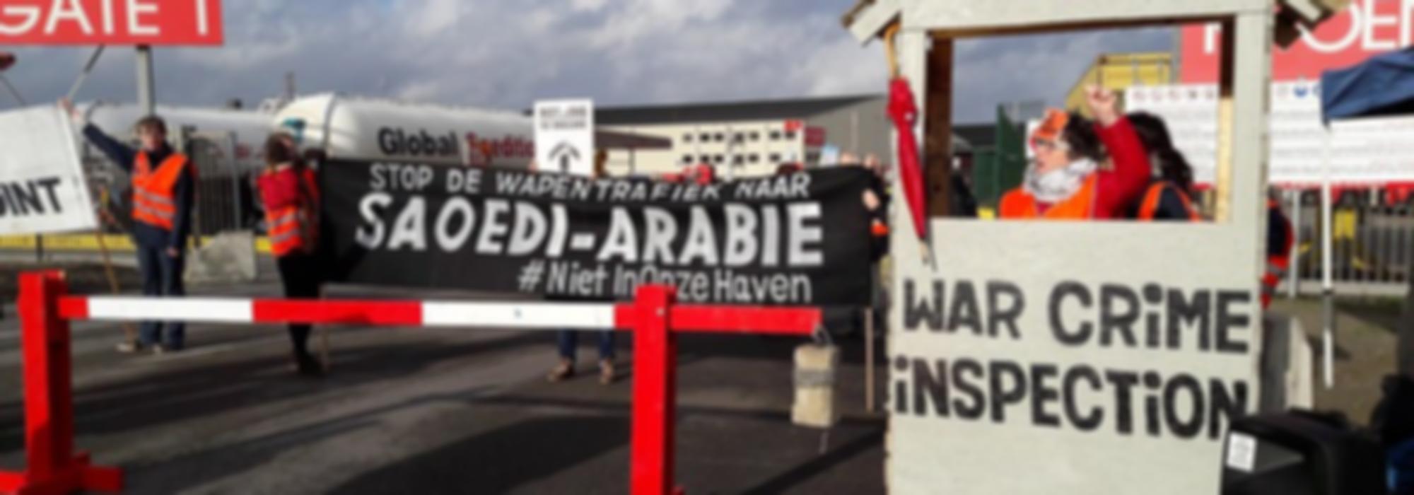 foto van bij protest aan de haven. We zien een zwarte banner met witte letters "Stop de wapenexport naar Saoedi-Arabië" en een vrouw in een douanehokje waarop "War Crime Inspectors" staat die haar vuist opsteekt