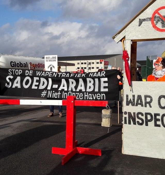 foto van bij protest aan de haven. We zien een zwarte banner met witte letters "Stop de wapenexport naar Saoedi-Arabië" en een vrouw in een douanehokje waarop "War Crime Inspectors" staat die haar vuist opsteekt